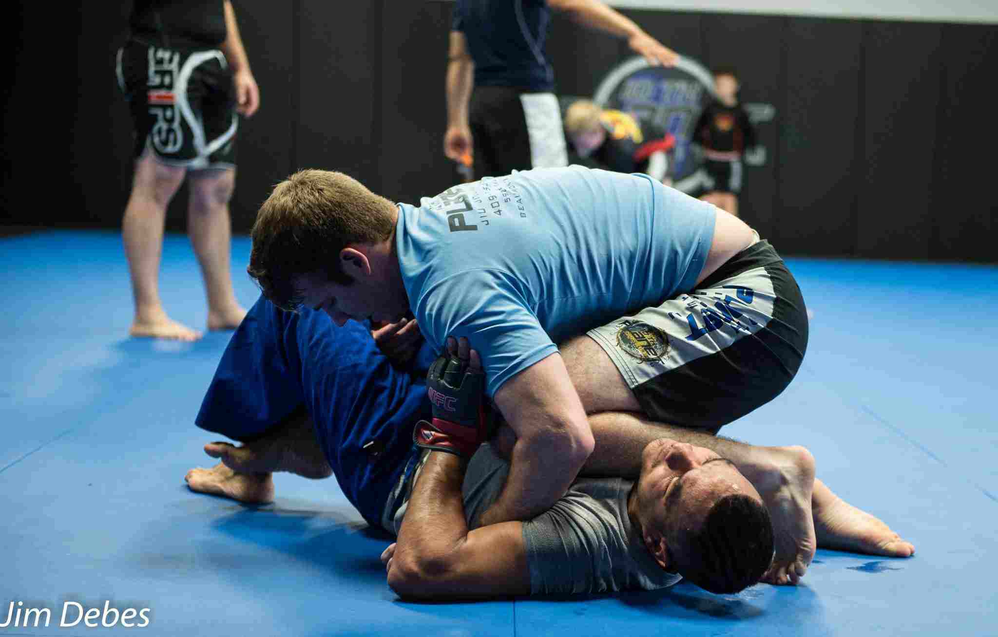 Side view of a 10th Planet blue belt adult male in a Brazilian jiu-jitsu class kneeling on another adult male.