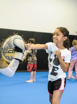 A ponytailed girl in the foreground with a boy and a girl in the background are learning proper punching techniques.